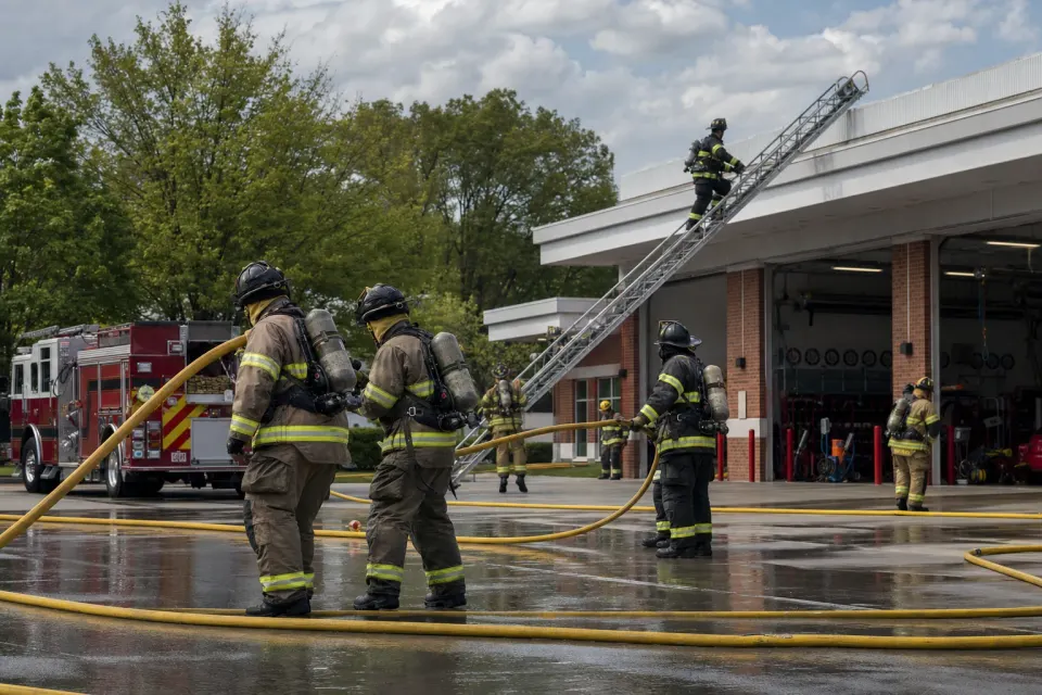 Firefighters handling hoses and a ladder during training outside a fire station