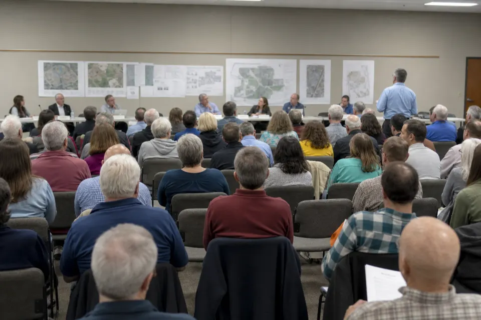 Residents seated in a public meeting room facing a panel and wall maps